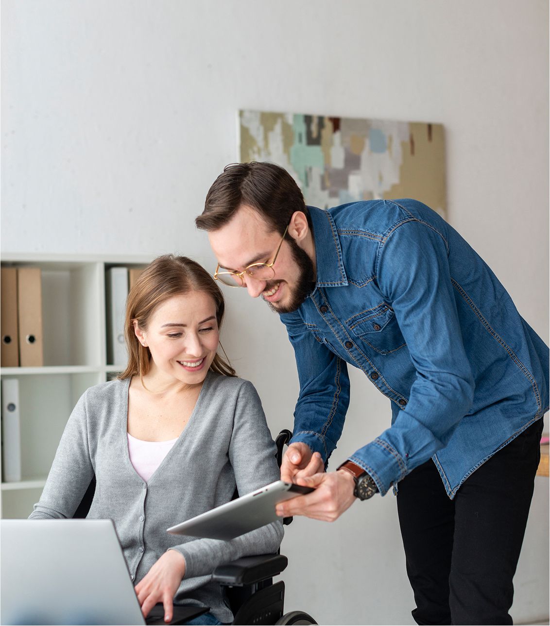 A support worker shows a tablet to a smiling woman using a wheelchair as they work together at a desk