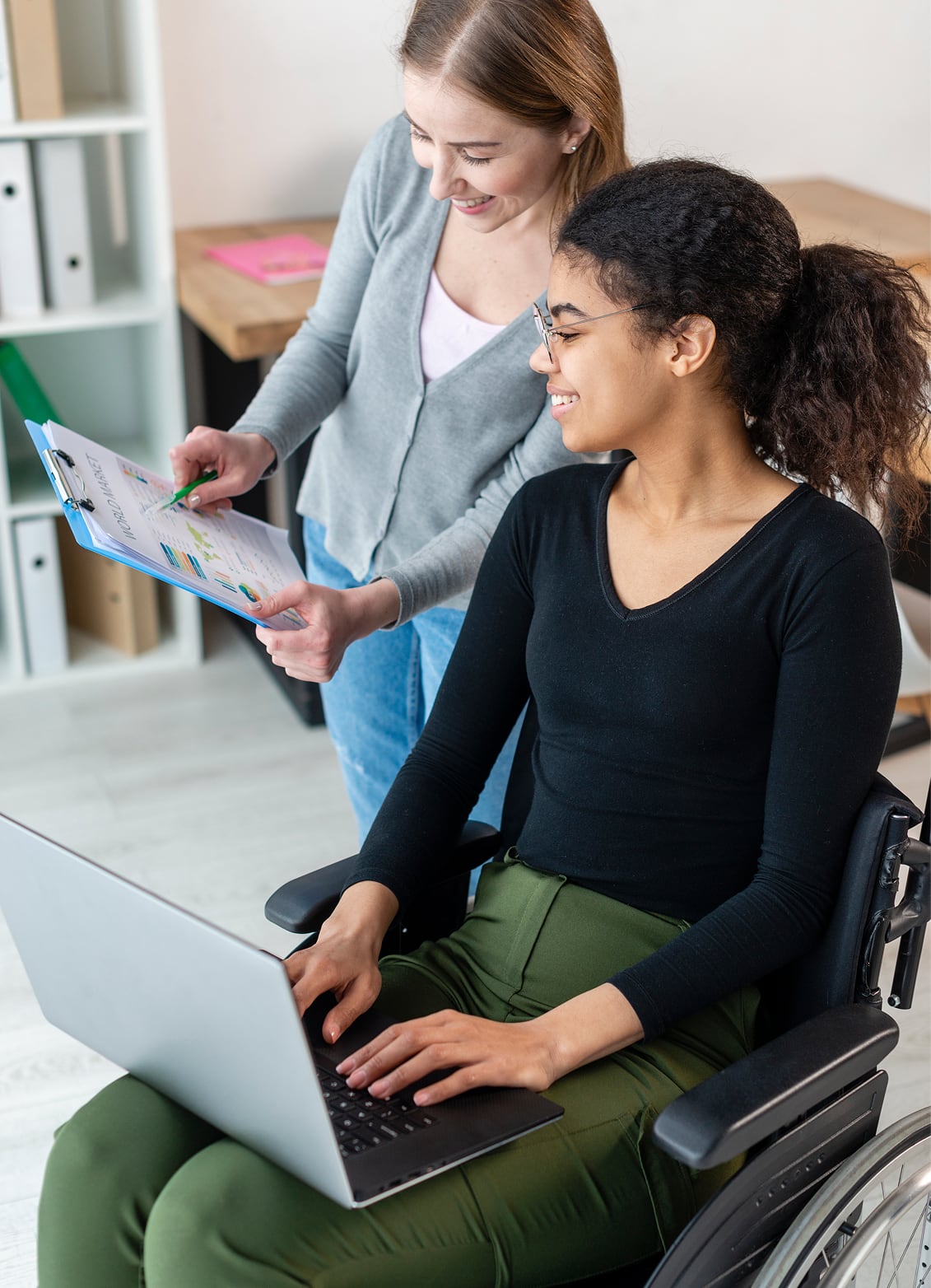 A worker points at a clipboard while a young woman in a wheelchair types on a laptop
