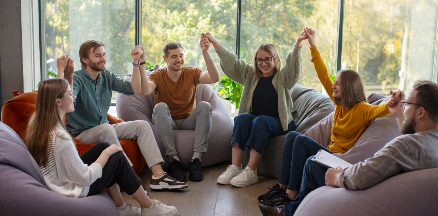 A support group of seven people celebrating together, holding hands raised in a sunlit room