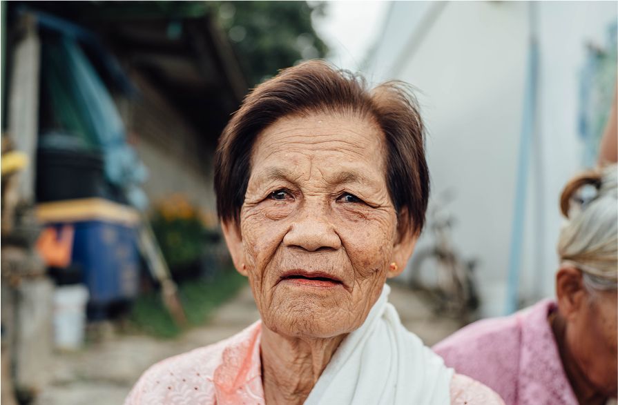 An older woman looks calmly at the camera outside her home
