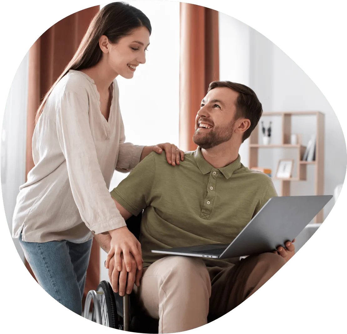A support worker and a man using a wheelchair share a laugh over a laptop at home