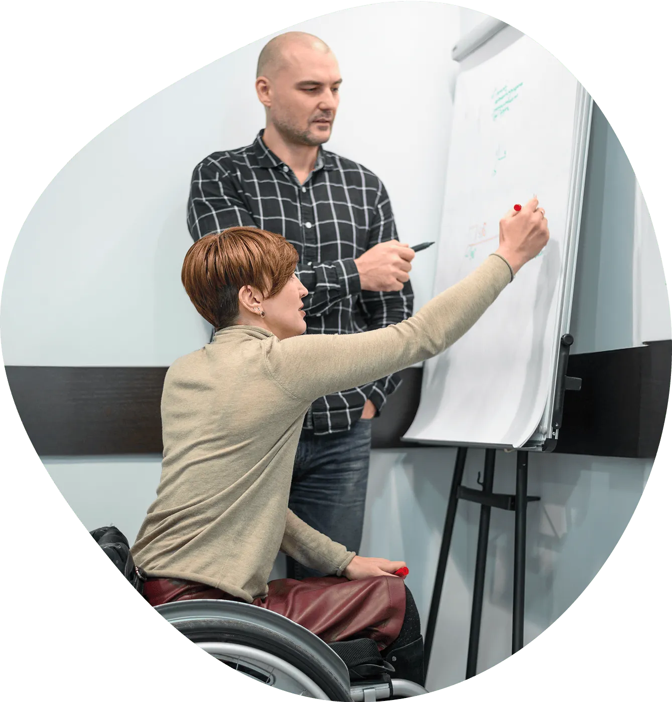 A woman in a wheelchair writes on a whiteboard while a colleague looks on
